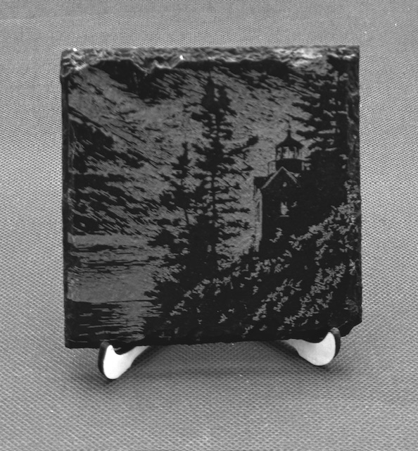 Black and white painting of a house on a hill near a lake under a cloudy sky, mounted on a stand.
