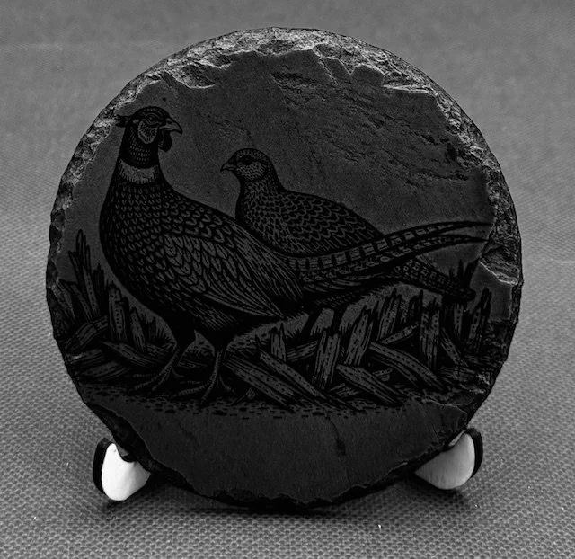 Black oval slate with engraved image of a quail or partridge, resting on branches with plants in the background.