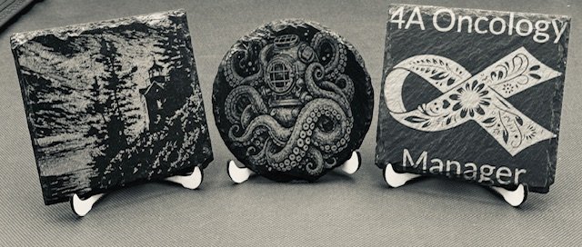 Three small display stands holding stones engraved with artwork, including a forest scene, an octopus design, and an oncology manager logo.