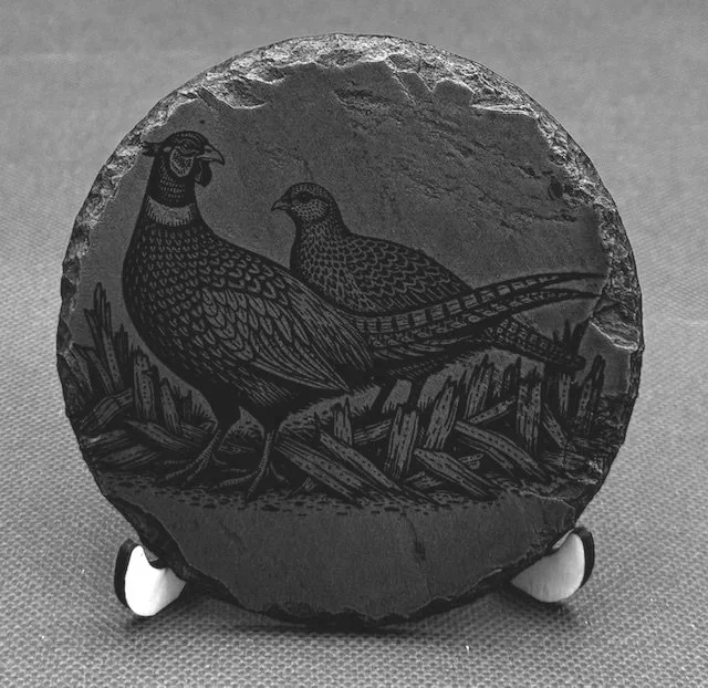 Black and light grey engraved image of a grouse and a partridge, with grass and logs in the background, on a round stone plaque displayed on a stand.