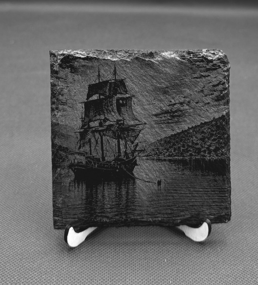 A small square artwork depicting a sailing ship on calm water with a rolling hillside in the background, mounted on a white stand.