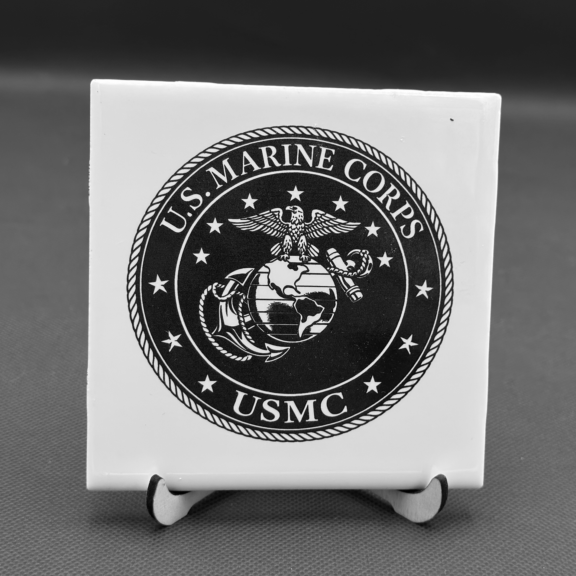 White plaque engraved  with a black-and-white U.S. Marine Corps emblem on a rectangular object, placed on a small stand with rolled edges, against a textured surface background.