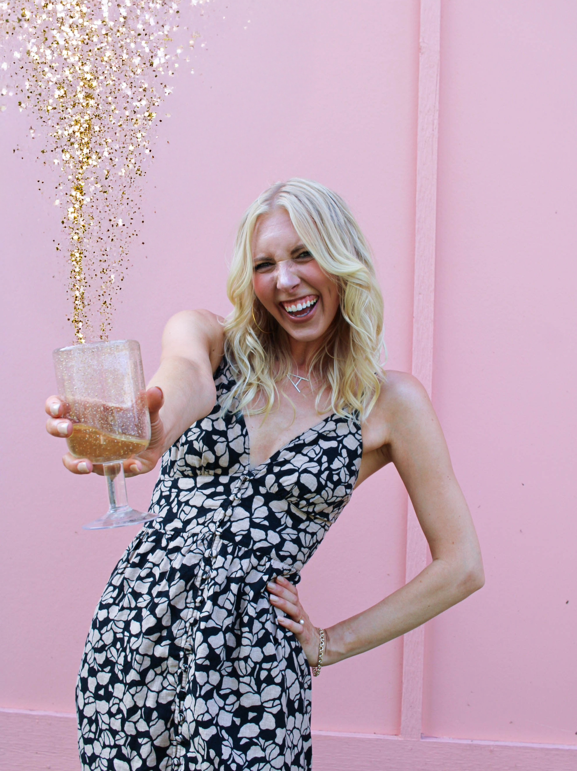 A smiling blonde woman in a black and white patterned dress holding a glass of sparkling wine against a pink wall, with gold glitter spilling from the glass.