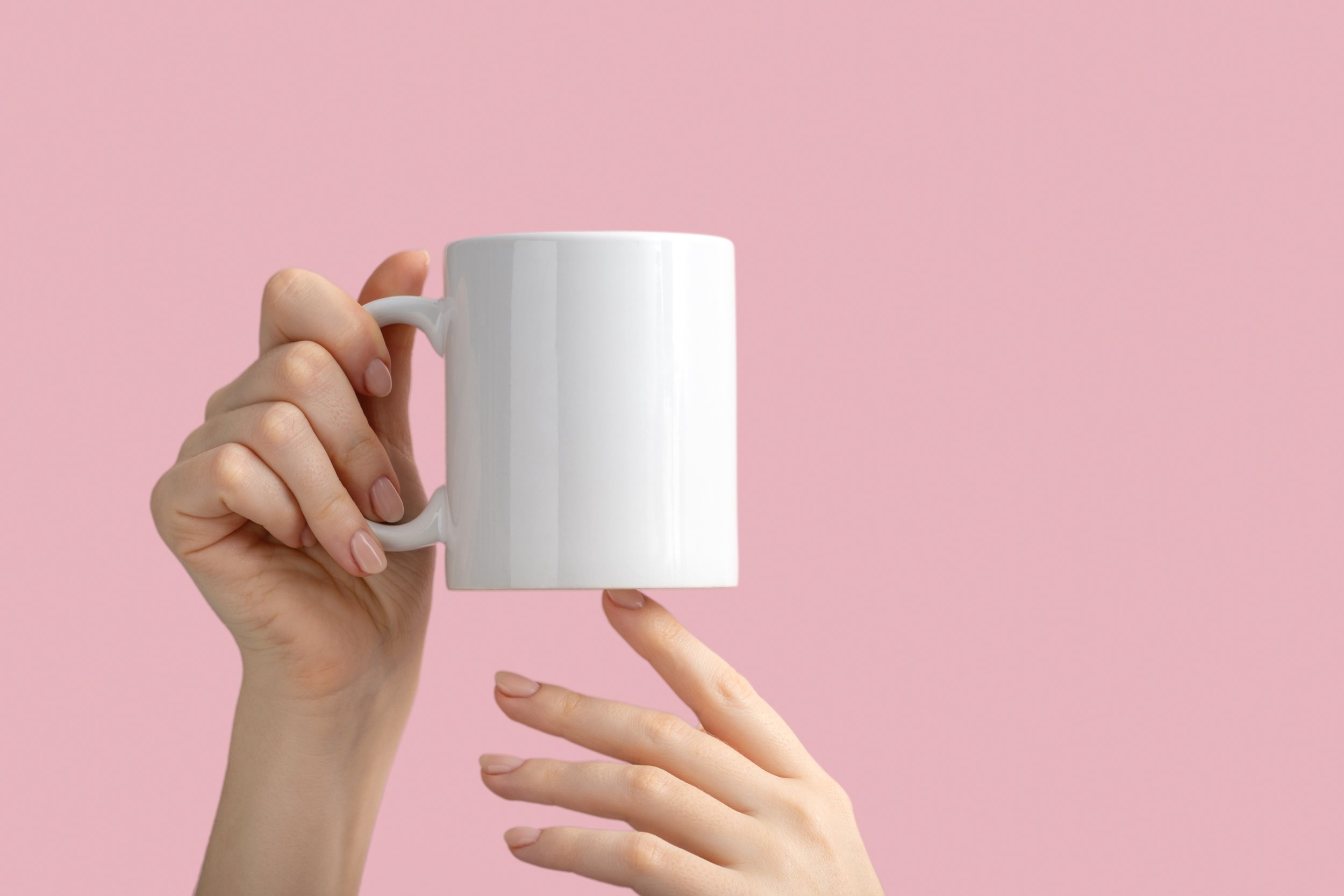 A person holding a white coffee mug against a pink background, with one hand and several fingers of the other hand visible pointing toward the mug.