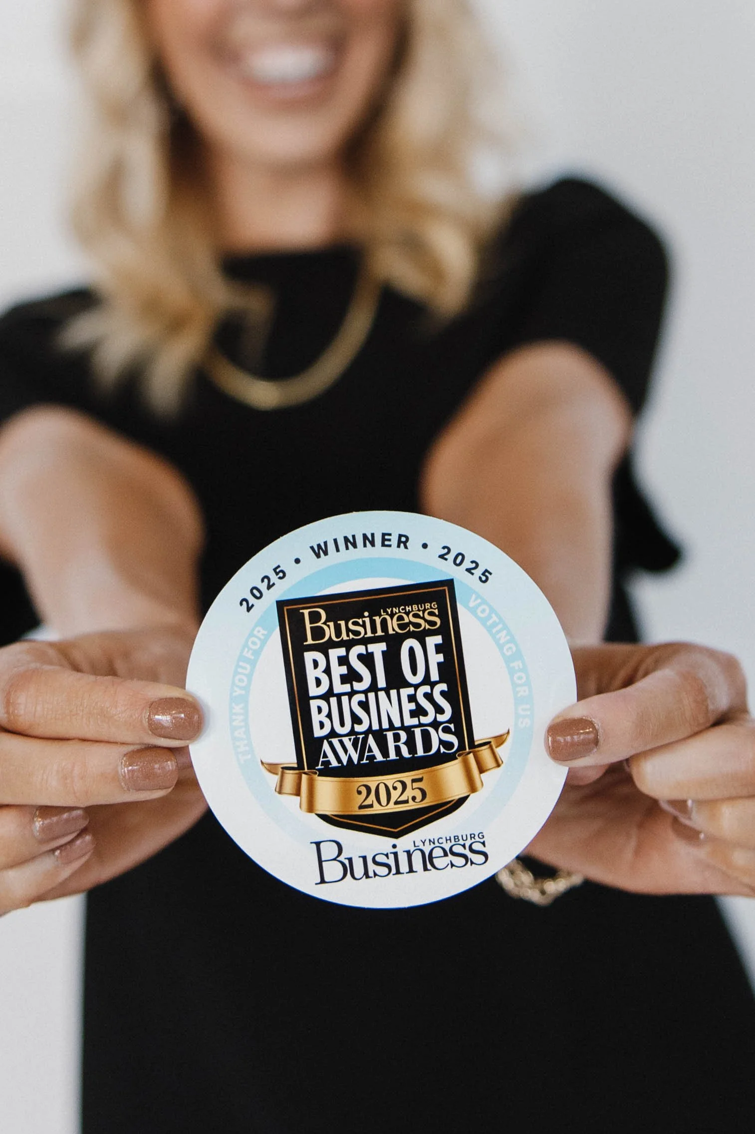 Close-up of a person holding a circular award sticker that says "Best of Business Awards 2025." The person has blonde hair, is smiling, and is wearing a black top.