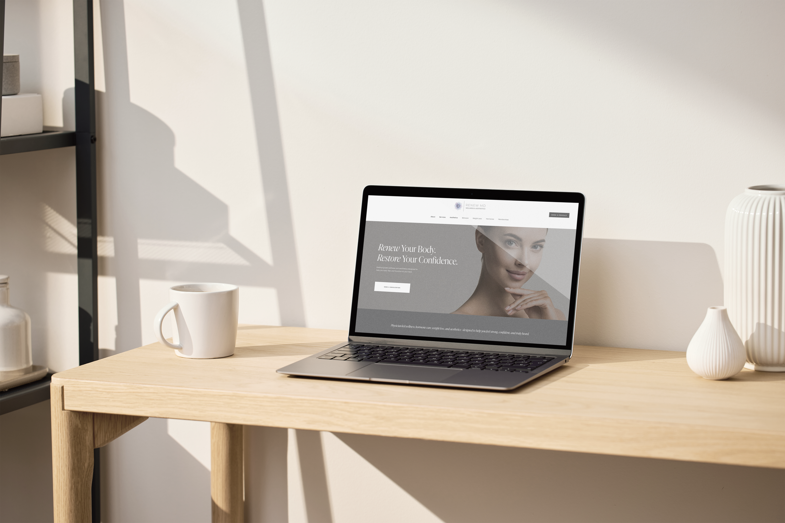 A wooden desk with a silver laptop displaying a website named 'Renew MD' featuring a woman's face, a white coffee mug, and decorative vases, with sunlight casting shadows on a light-colored wall.