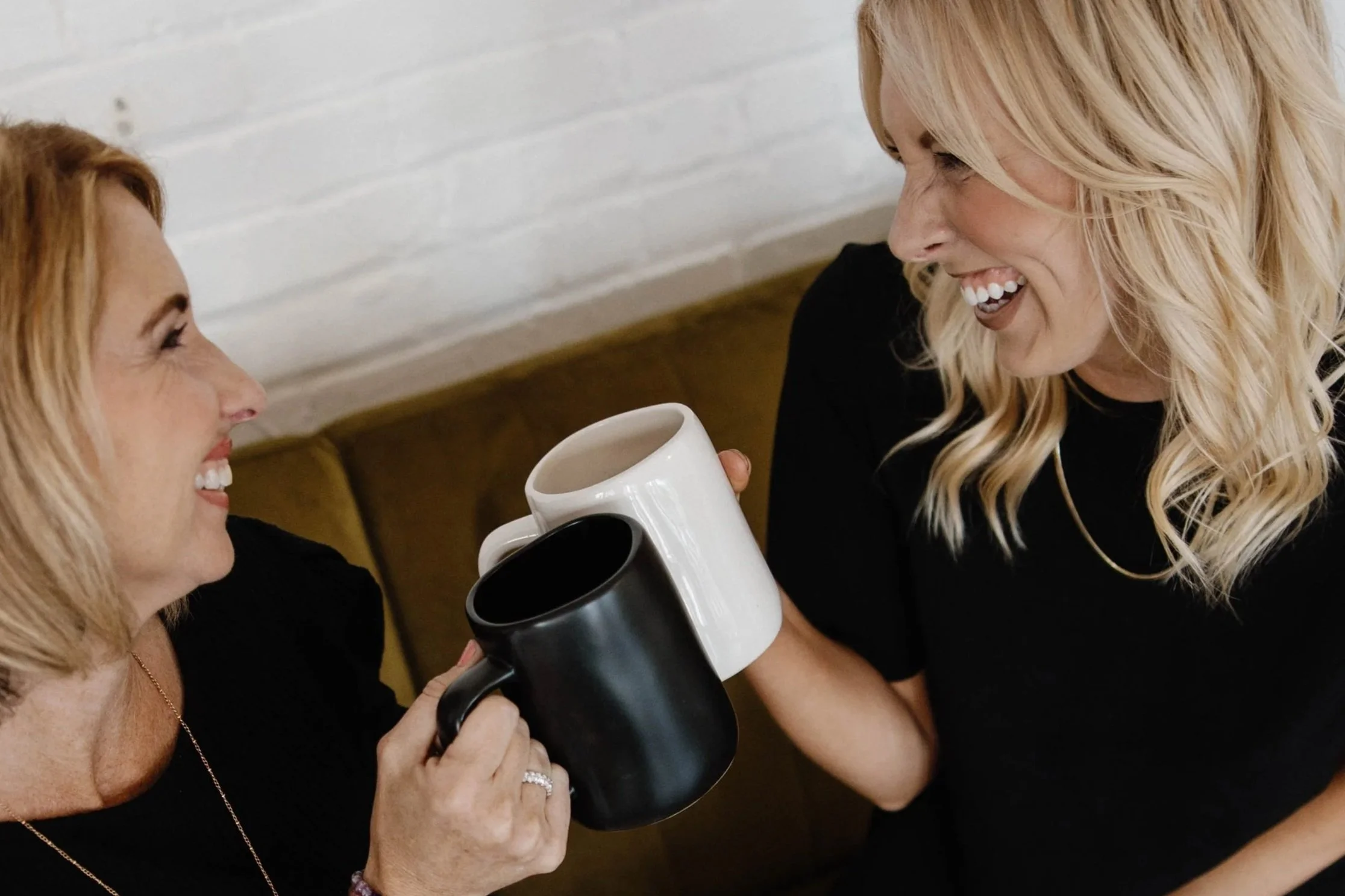 Two women smiling, one older with blonde hair holding a black coffee mug, and a younger woman with blonde hair, holding a white coffee mug, sharing a joyful moment.