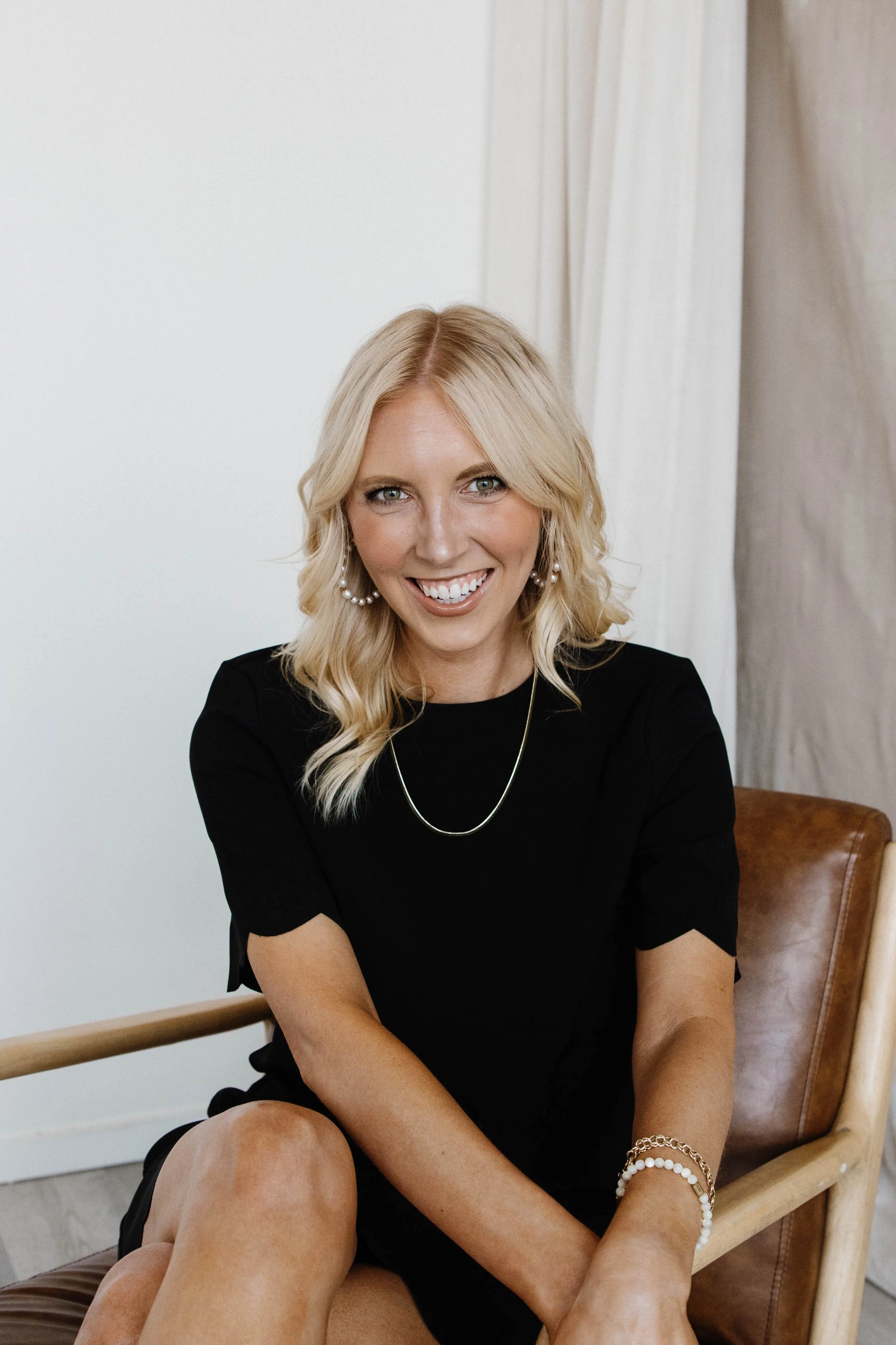 A woman with blonde hair, blue eyes, wearing a black dress, large pearl earrings, a pearl bracelet, and a gold chain necklace, sitting on a brown chair.