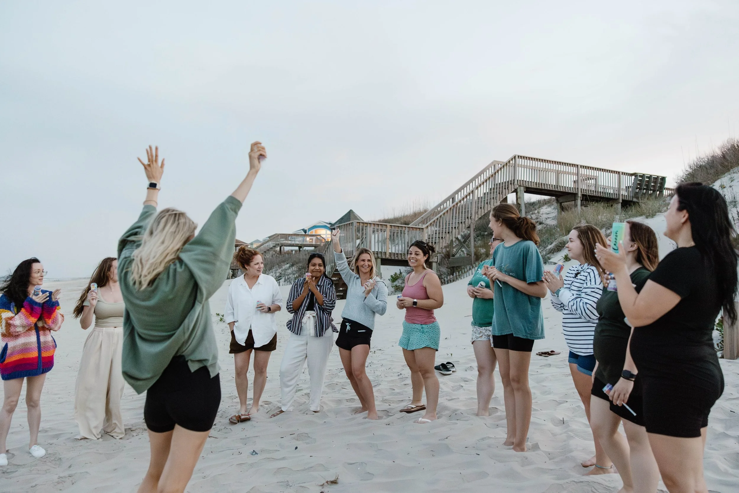 A group of women standing on the beach, smiling and clapping while a woman in green gestures with her hands. Some women hold water bottles, and a wooden staircase and beach house are visible in the background during late afternoon or early evening.