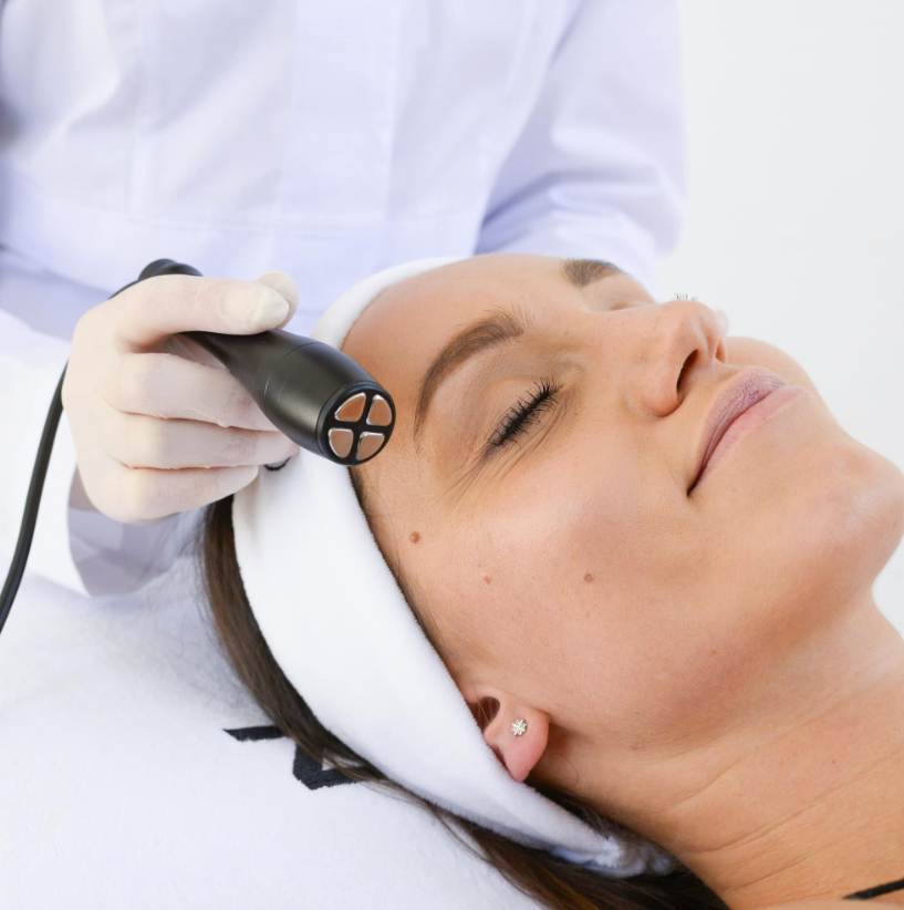 A woman receiving a facial treatment with a handheld device, lying on a treatment bed with her eyes closed, wearing a white headband and diamond-shaped stud earrings.