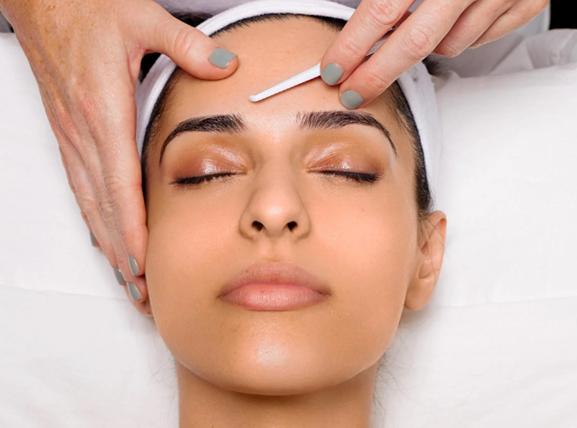 A woman receiving a facial treatment, with her eyes closed, as a technician uses a tool on her forehead.