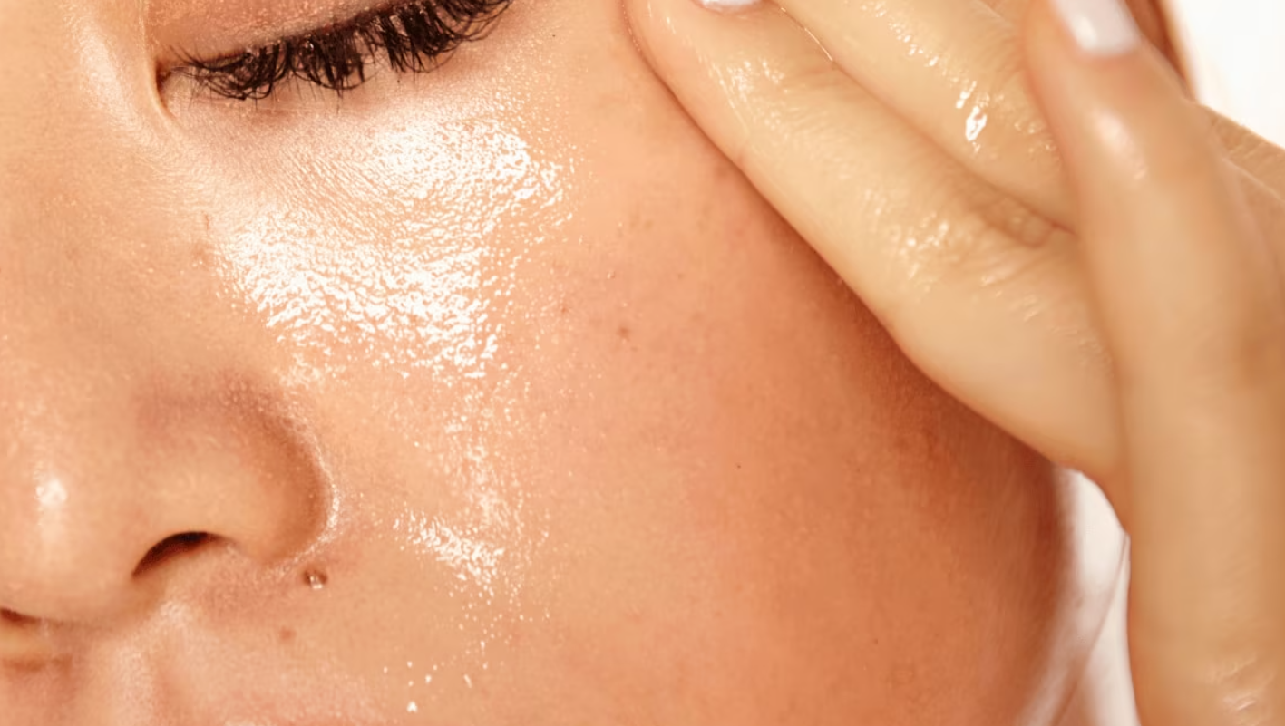 Close-up of a person’s face and hand with shiny, moisturized skin, eye closed, and fingers gently touching the cheek.