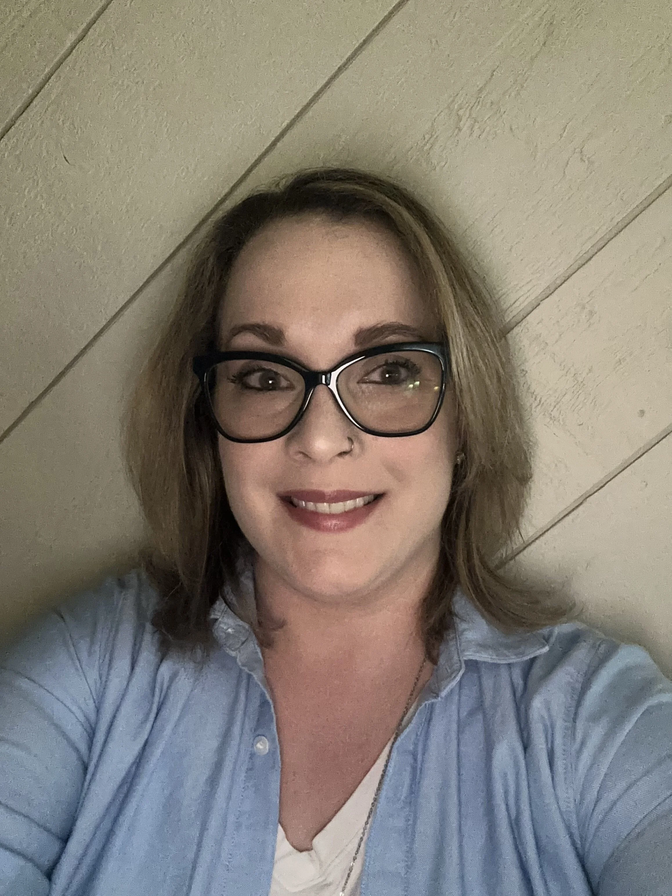 A woman with shoulder-length light brown hair, glasses, and fair skin smiling at the camera. She is wearing a light blue shirt over a white top and standing against a wood-paneled wall.