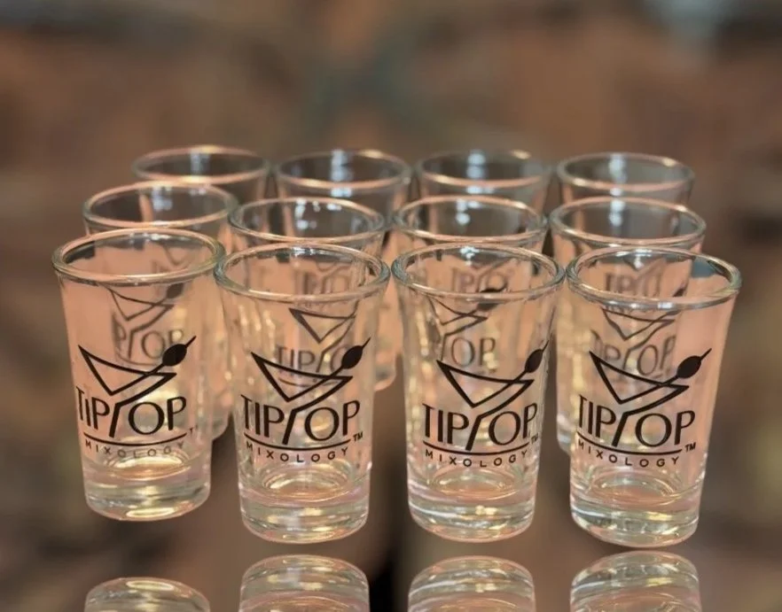 A group of 10 shot glasses with TIPOP Mixology logo on them arranged on a reflective surface.
