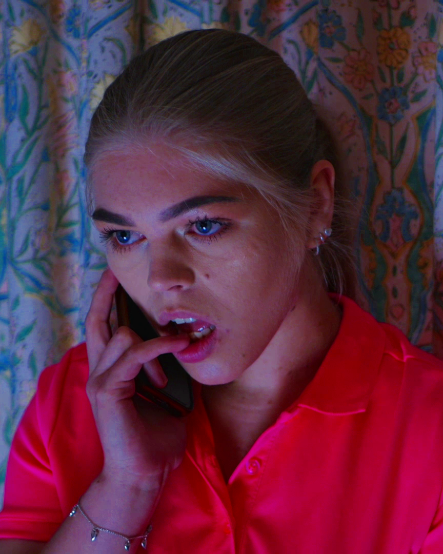 A woman with blonde hair and blue eyes wearing a pink shirt is speaking on the phone, with a colorful patterned curtain in the background.