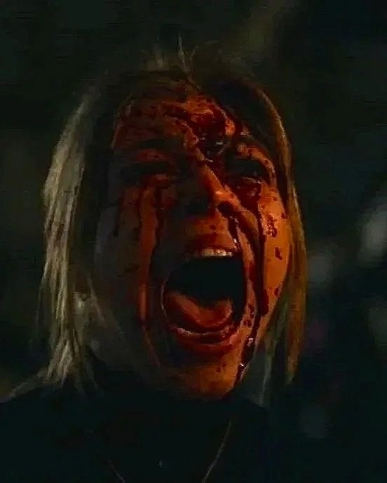 A person with long hair has blood streaming down their face, mouth open in a scream, in a dark setting.
