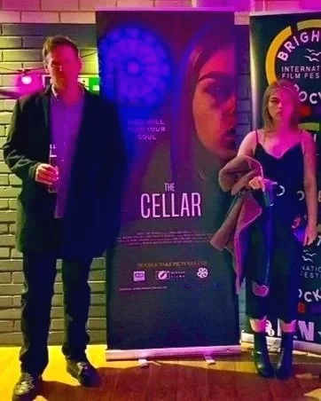 Two people standing in front of a poster for the film "The Cellar" at a film festival event. The person on the left is holding a drink and wearing a dark jacket, while the person on the right is holding a bag and wearing a dark dress.