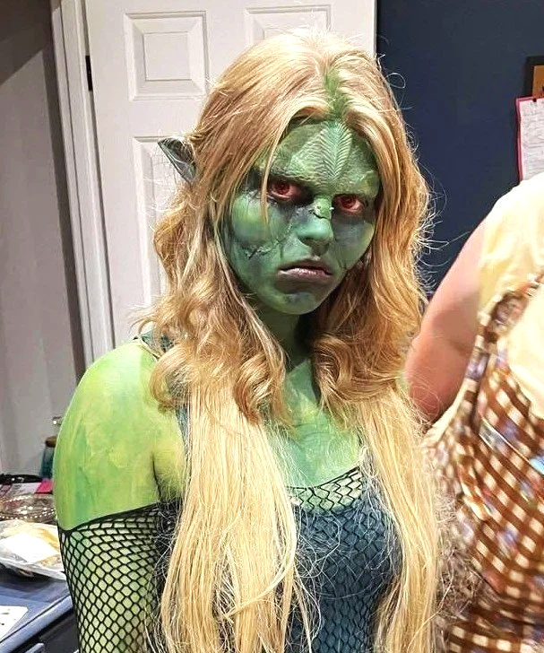 Person in detailed costume with green face paint, red eyes, long blonde hair, wearing a black patterned outfit.