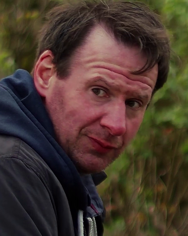 A man with short brown hair and light skin, wearing a dark jacket and hoodie, looking over his shoulder outside with greenery in the background.