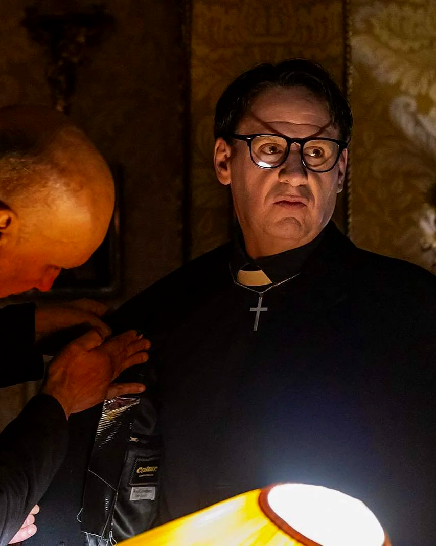A priest with glasses and a cross necklace looking surprised or upset while someone else touches his shoulder in a dimly lit room.
