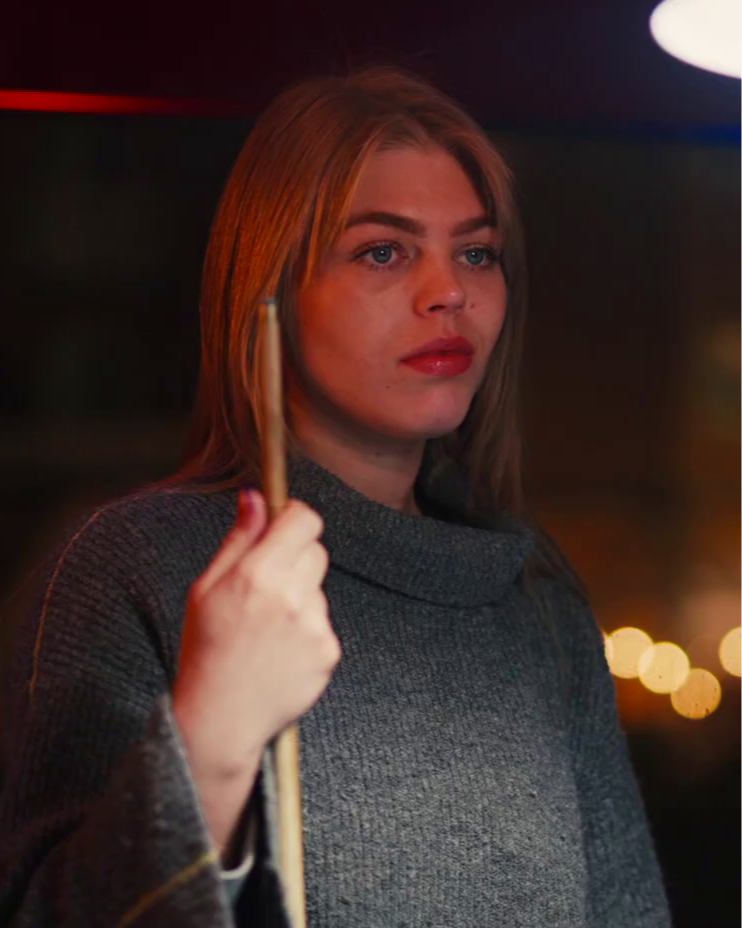 A young woman with blonde hair and blue eyes, wearing a gray sweater, holds a pool cue and looks thoughtfully into the distance, with blurred lights in the background.