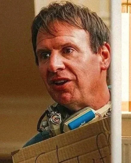 A man with brown hair talking, wearing a blue shirt, holding a cardboard box with an object inside, and standing behind a white pole.