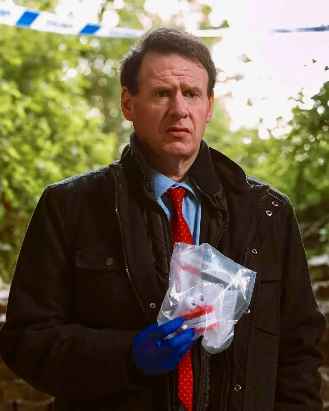 A man in a black jacket holding a plastic bag with a clown face in an outdoor setting with trees in the background.