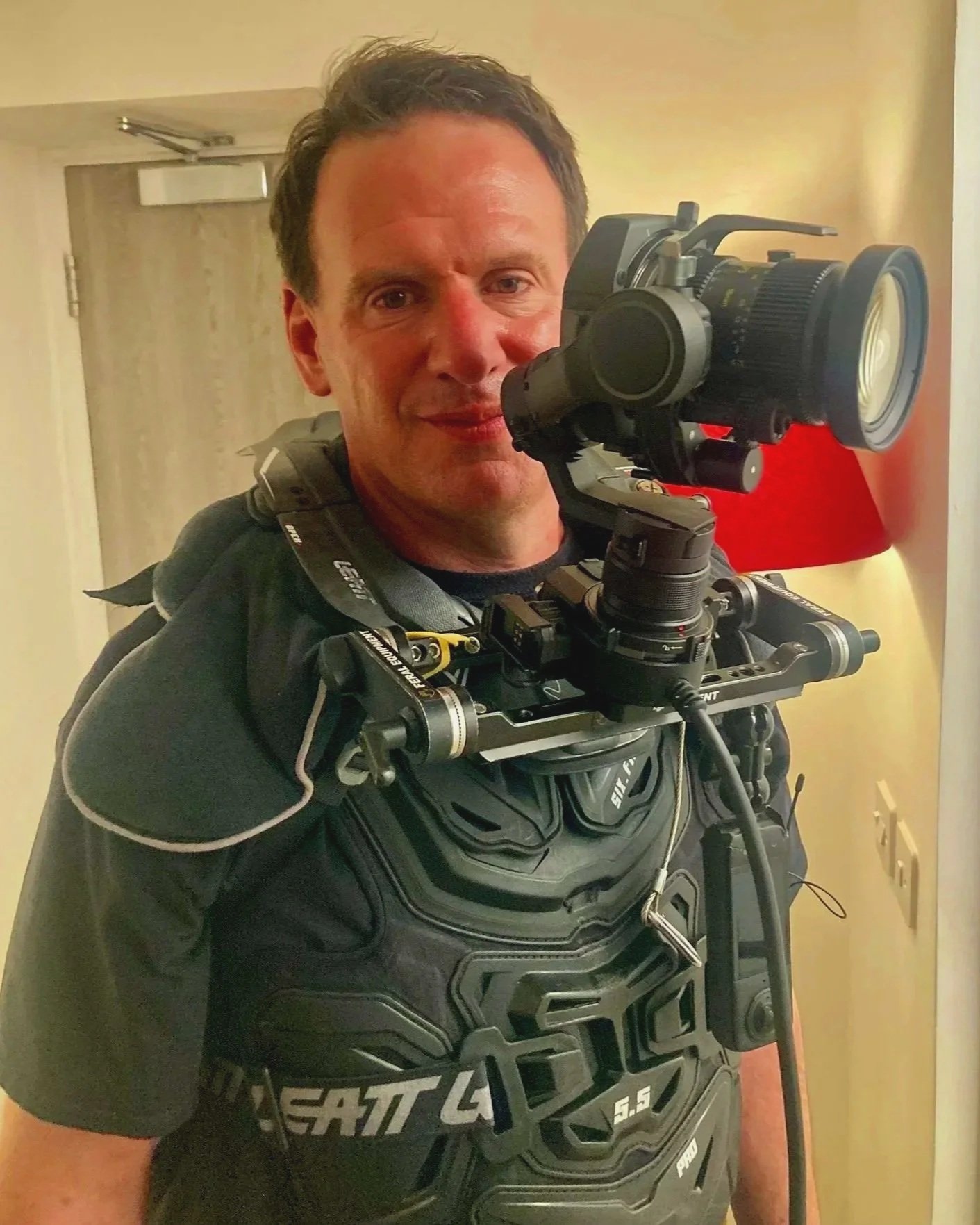 A man wearing a motorcycle jacket and chest protector is taking a selfie using a camera mounted on a stabilizer, standing in a hallway with a door behind him.