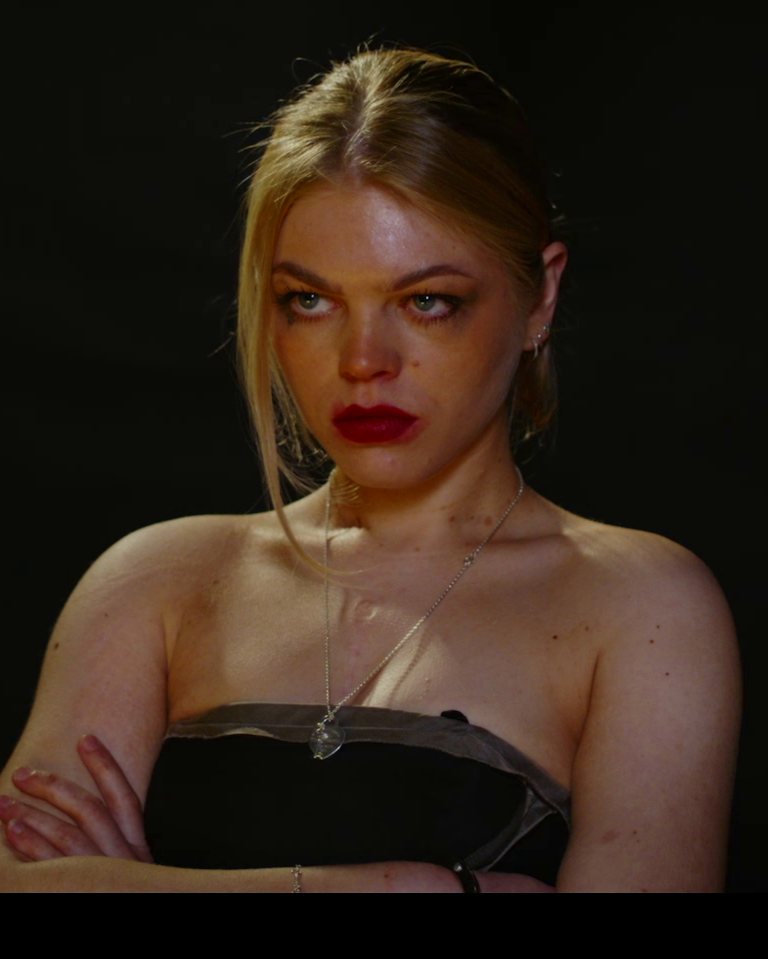 A woman with blonde hair, red lipstick, and a serious expression, wearing a strapless top and a necklace, crossing her arms against a dark background.