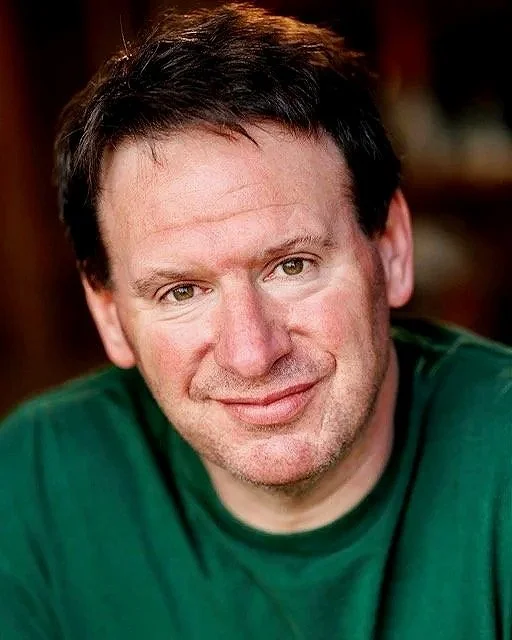 A smiling man with dark hair wearing a green shirt in a warmly lit indoor setting.
