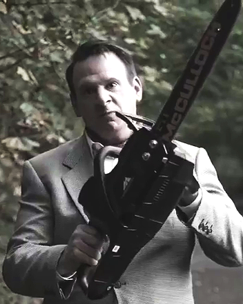 A man in a suit holding a chainsaw in front of a wooded background.