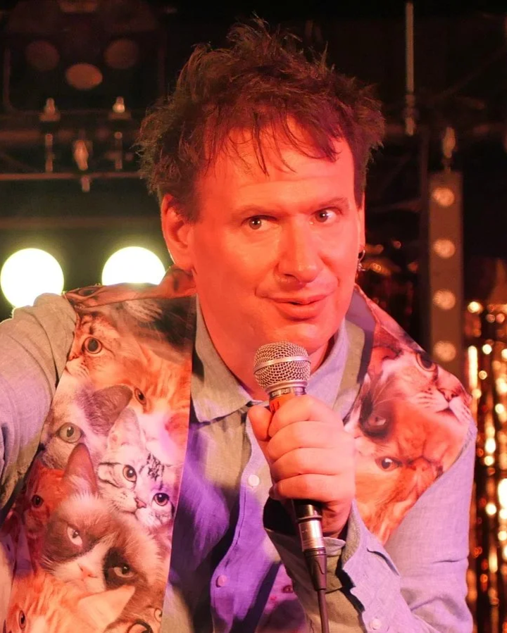 A man with messy brown hair holding a microphone, wearing a shirt with a vest covered in various cat images, standing on a stage with bright lights and a dark background.