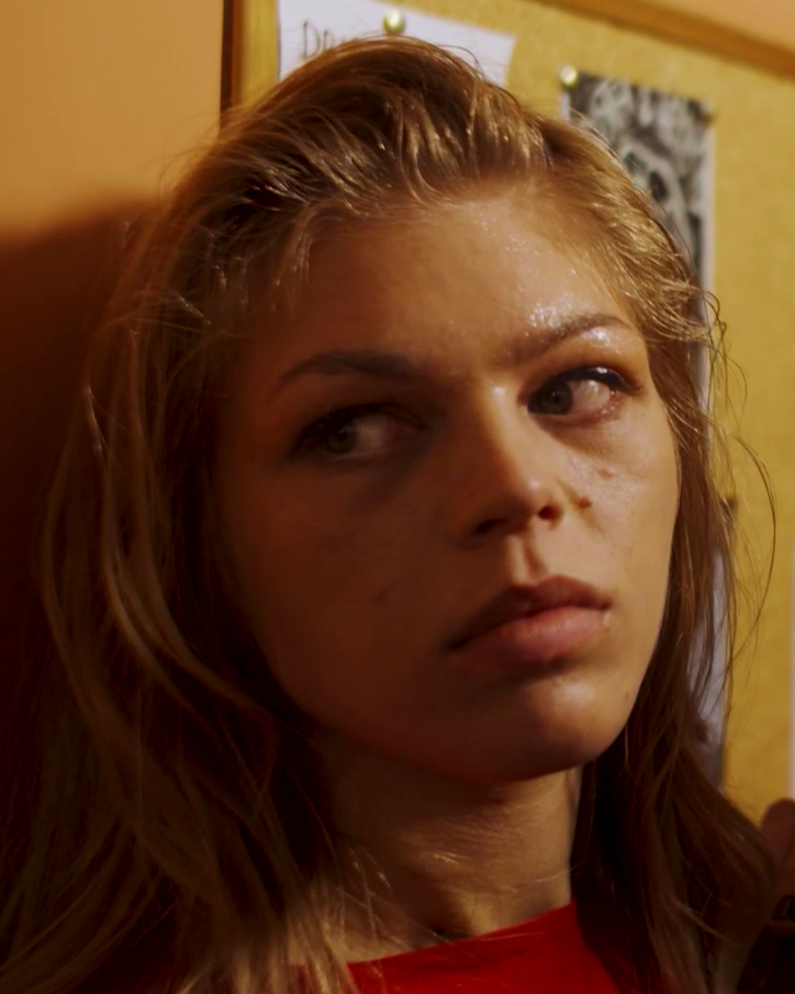A young woman with light brown hair and a serious expression, standing against a yellow wall with posters pinned on cork boards.