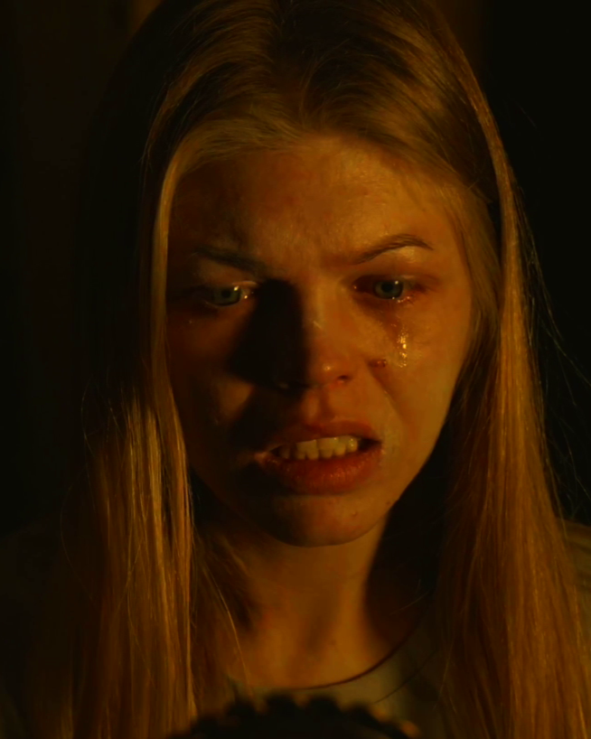 A young woman with long blonde hair appears distressed, with tears on her face and an intense expression in low, dramatic lighting.