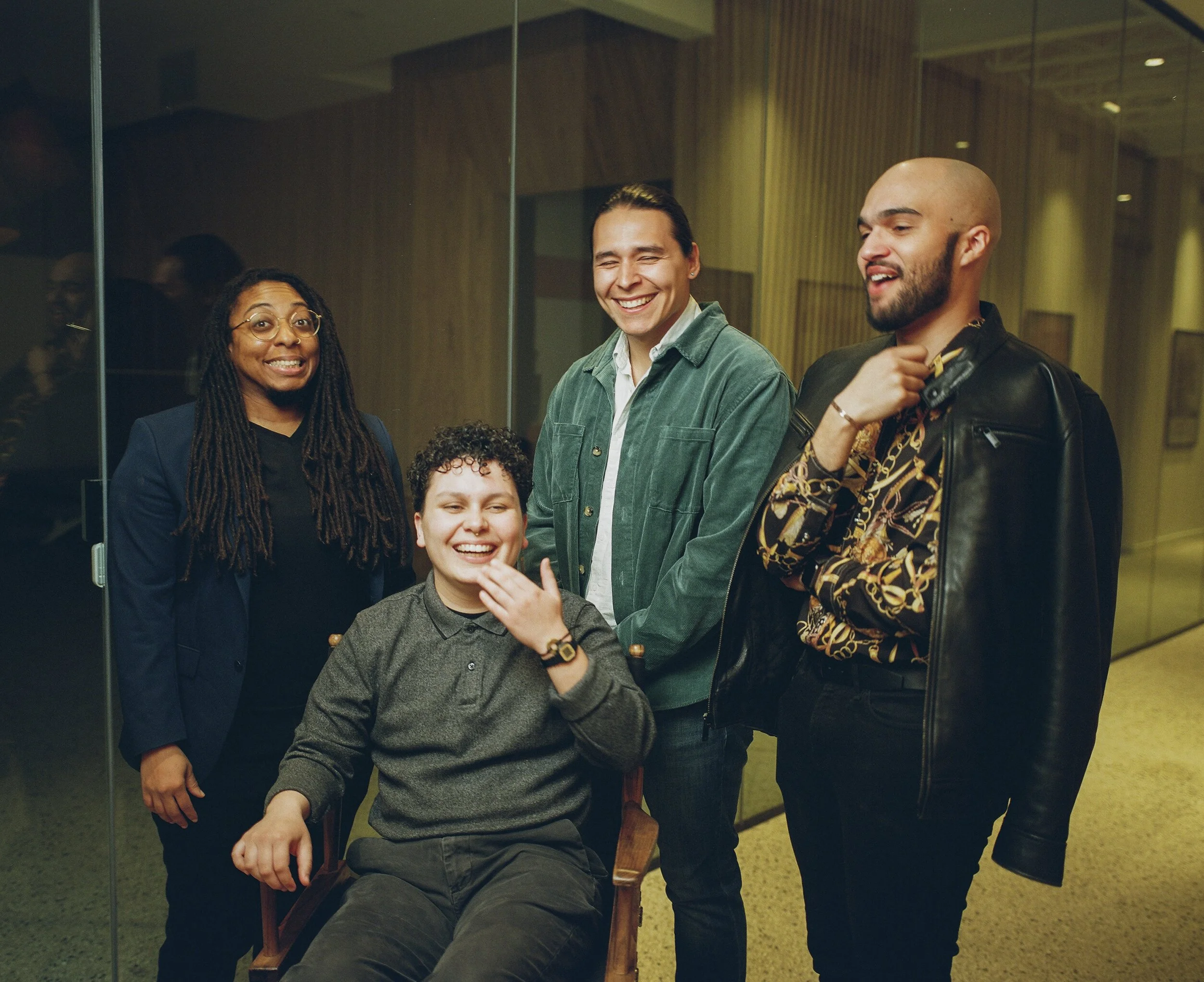 Group of four diverse smiling people, one sitting in a chair, three standing, engaged in conversation in an indoor setting.
