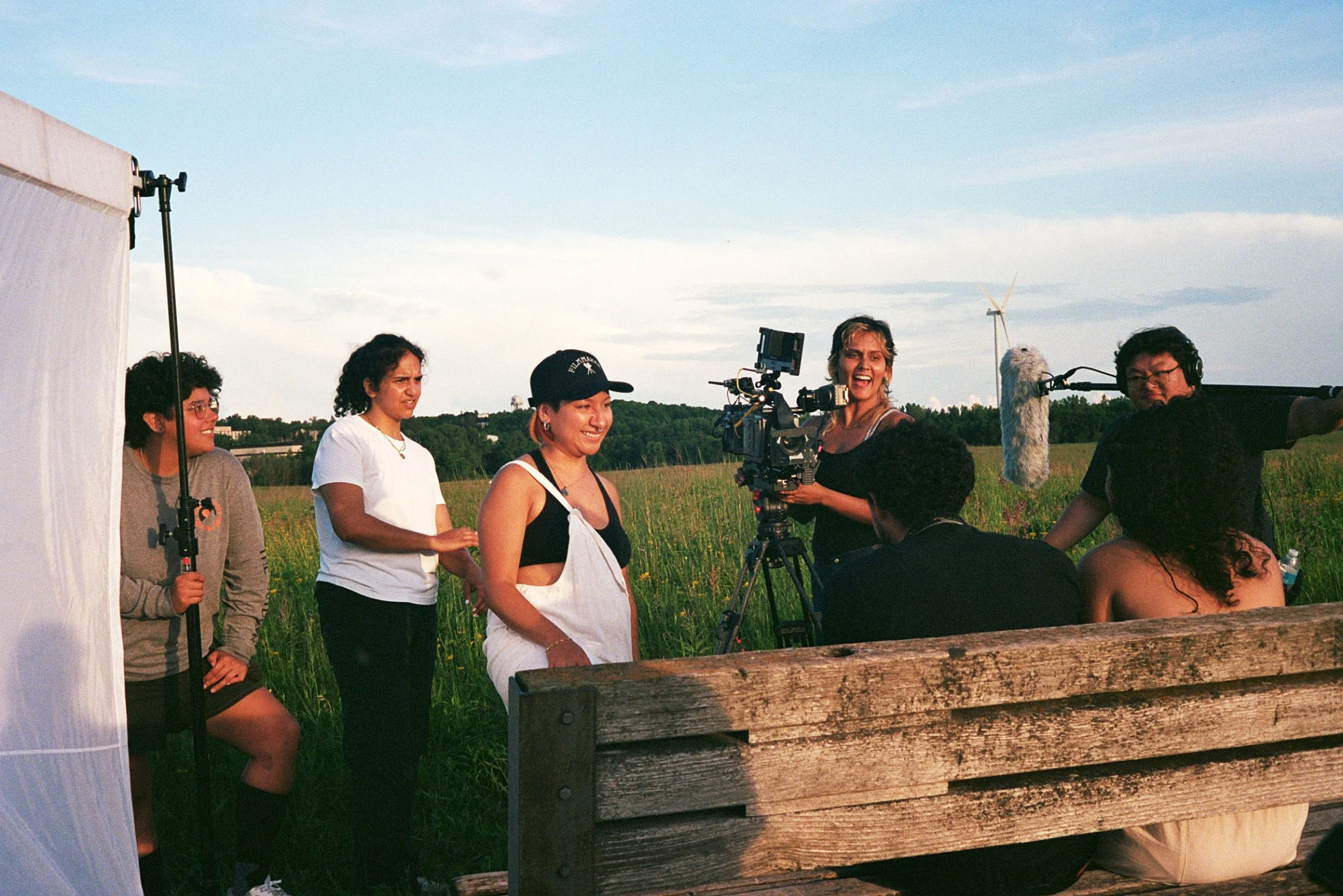 Filming crew capturing a scene outdoors in a grassy field during daylight with equipment such as a camera on a tripod, a large microphone, and reflectors, while crew members work and interact.