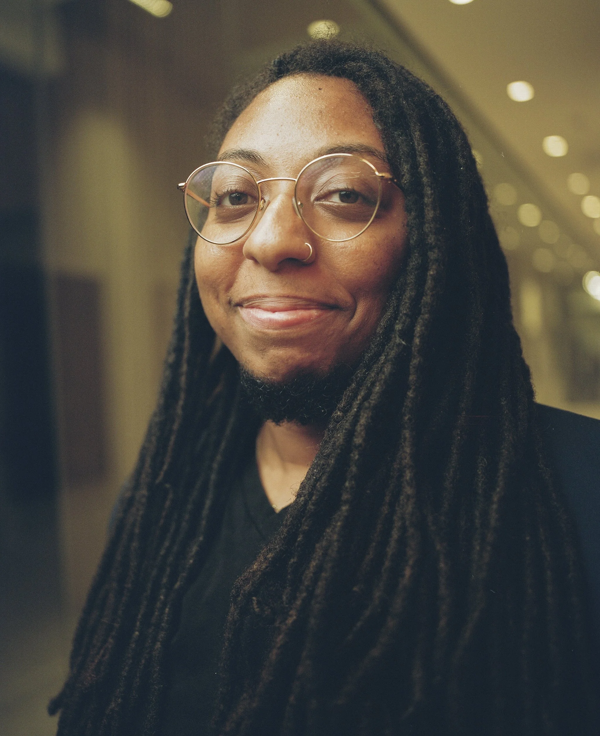 A woman with long dreadlocks, round glasses, and a nose ring smiling indoors.