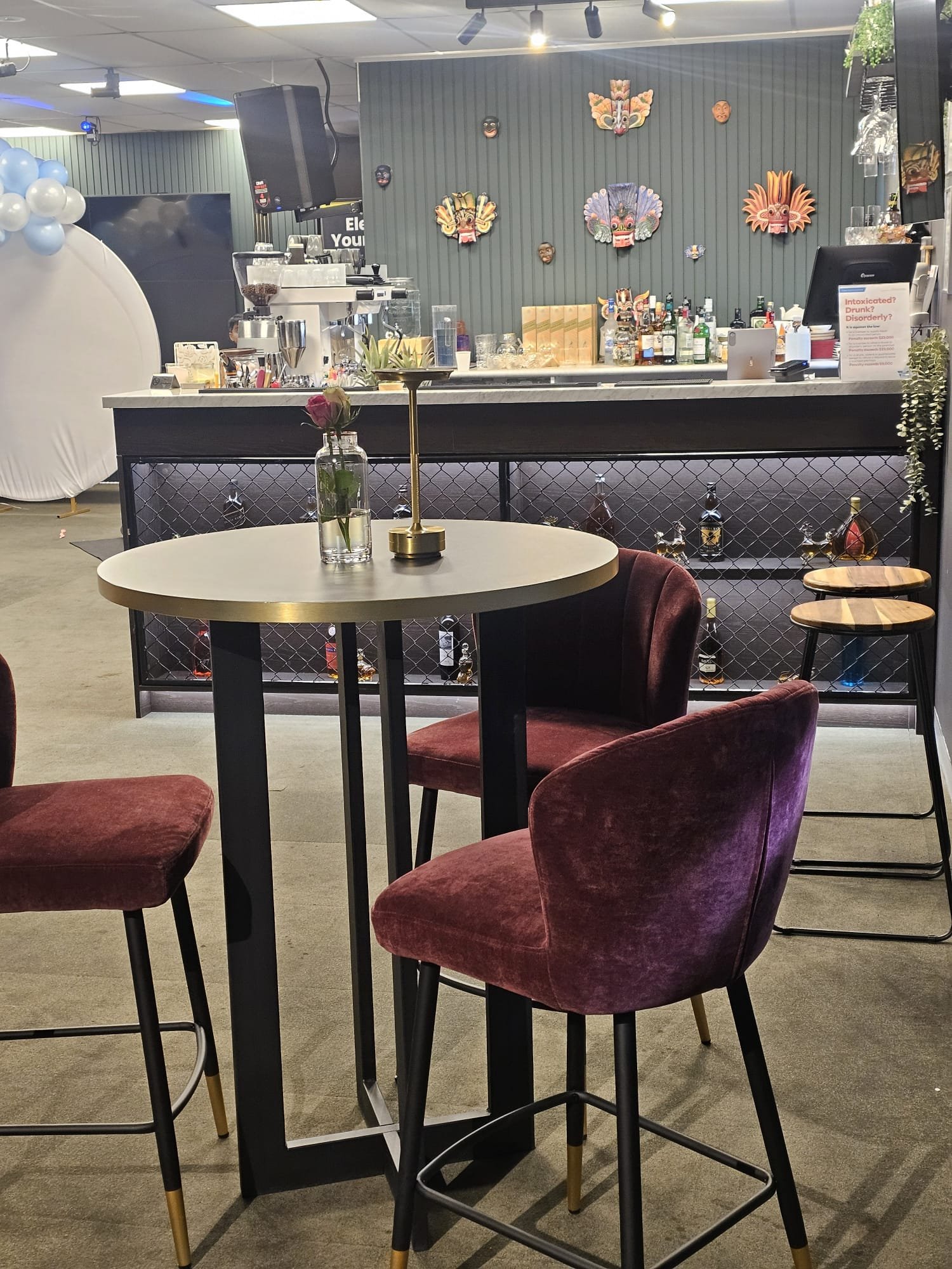 A cozy bar with a contemporary design, featuring a bar counter with bottles, glasses, and a coffee machine, decorated with colorful masks and artwork on the wall. There are tables and upholstered chairs in the foreground.