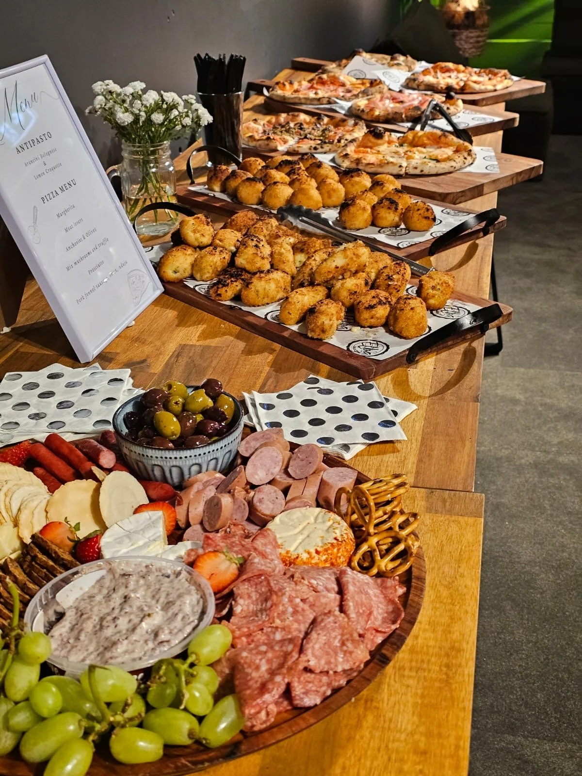 A buffet table with various appetizers including a charcuterie board with cured meats, strawberries, and cheese, a bowl of mixed green and purple olives, and several trays of pizza and fried mozzarella bites.