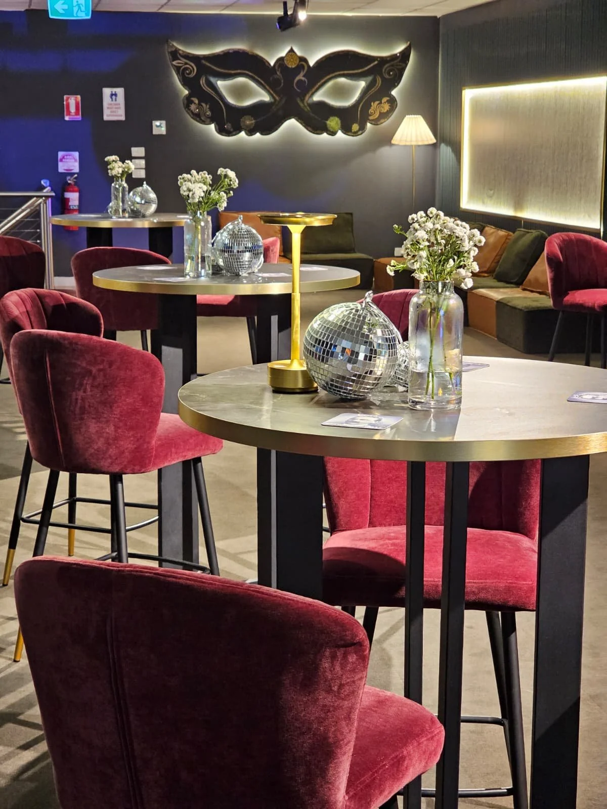 A stylish lounge with round tables, pink velvet chairs, floral arrangements, and disco ball decorations, illuminated by soft lighting and a large decorative wall piece in the shape of a mask.