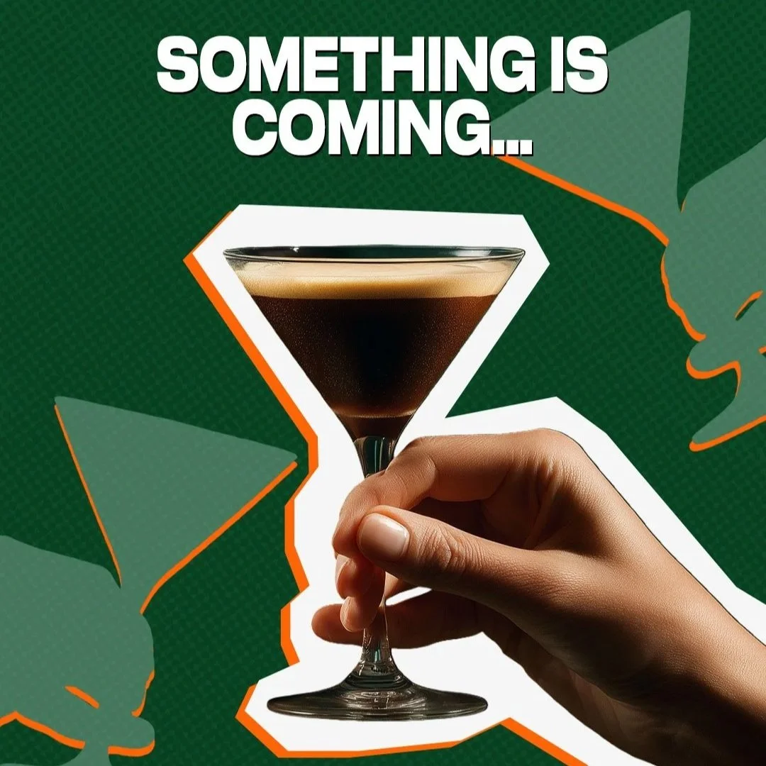A hand holding a martini glass filled with a dark beverage against a green background with tropical leaves and the text 'Something is coming...'