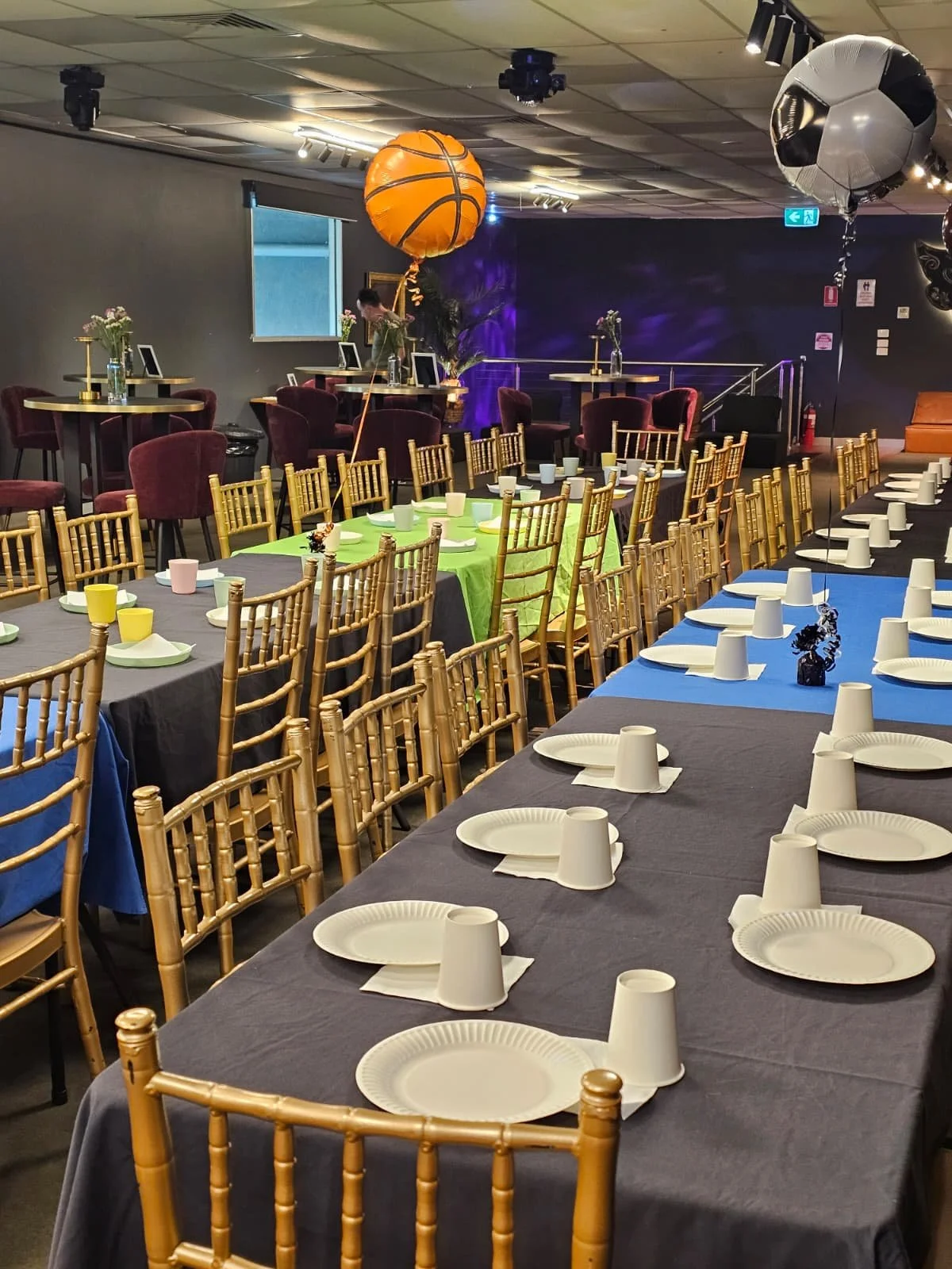 Event space with decorated tables, chairs, balloons including a basketball and a soccer ball, and table settings with paper plates, cups, and napkins.