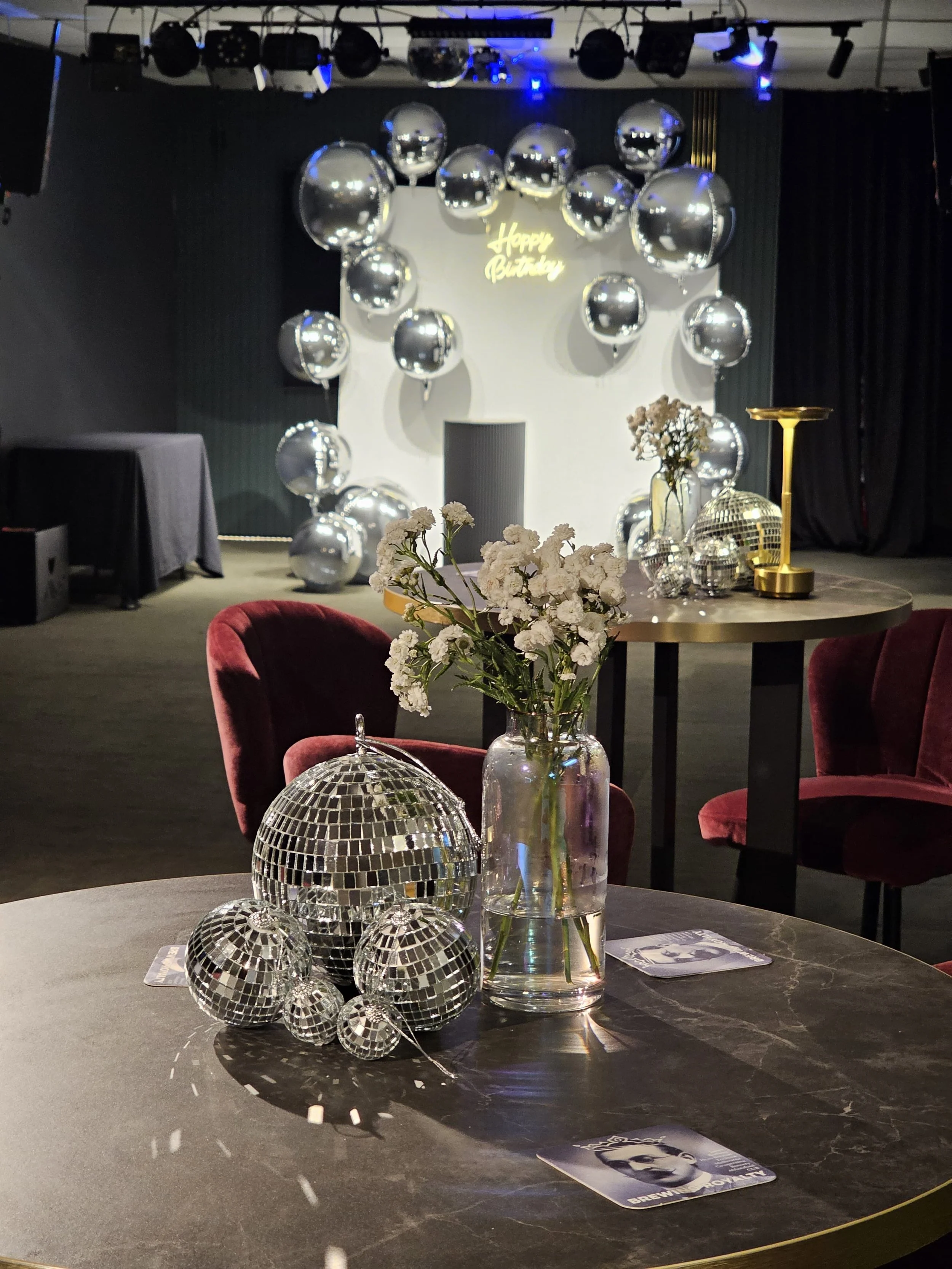 A decorated party setup with a backdrop of balloons reflecting light and a neon sign that says 'Happy Birthday.' The foreground features a table with a clear vase of white flowers, several disco ball decorations, and a card with black and white portr