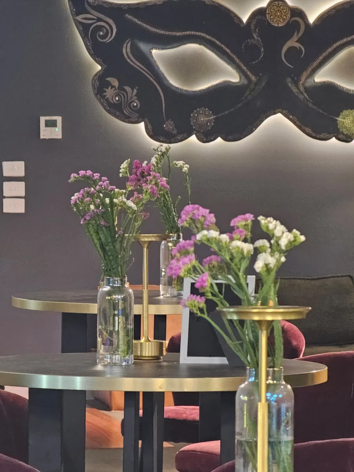 Interior of a modern dining space with round tables decorated with vases of pink and white flowers, set against a dark wall with a decorative mask and backlit lighting.