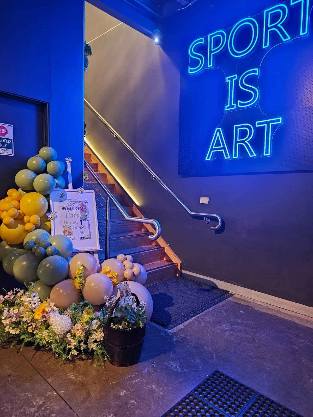 Entrance decorated with balloons and flowers, featuring a sign welcoming guests to a girl's birthday party, and a neon sign reading 'SPORT IS ART' on the wall.