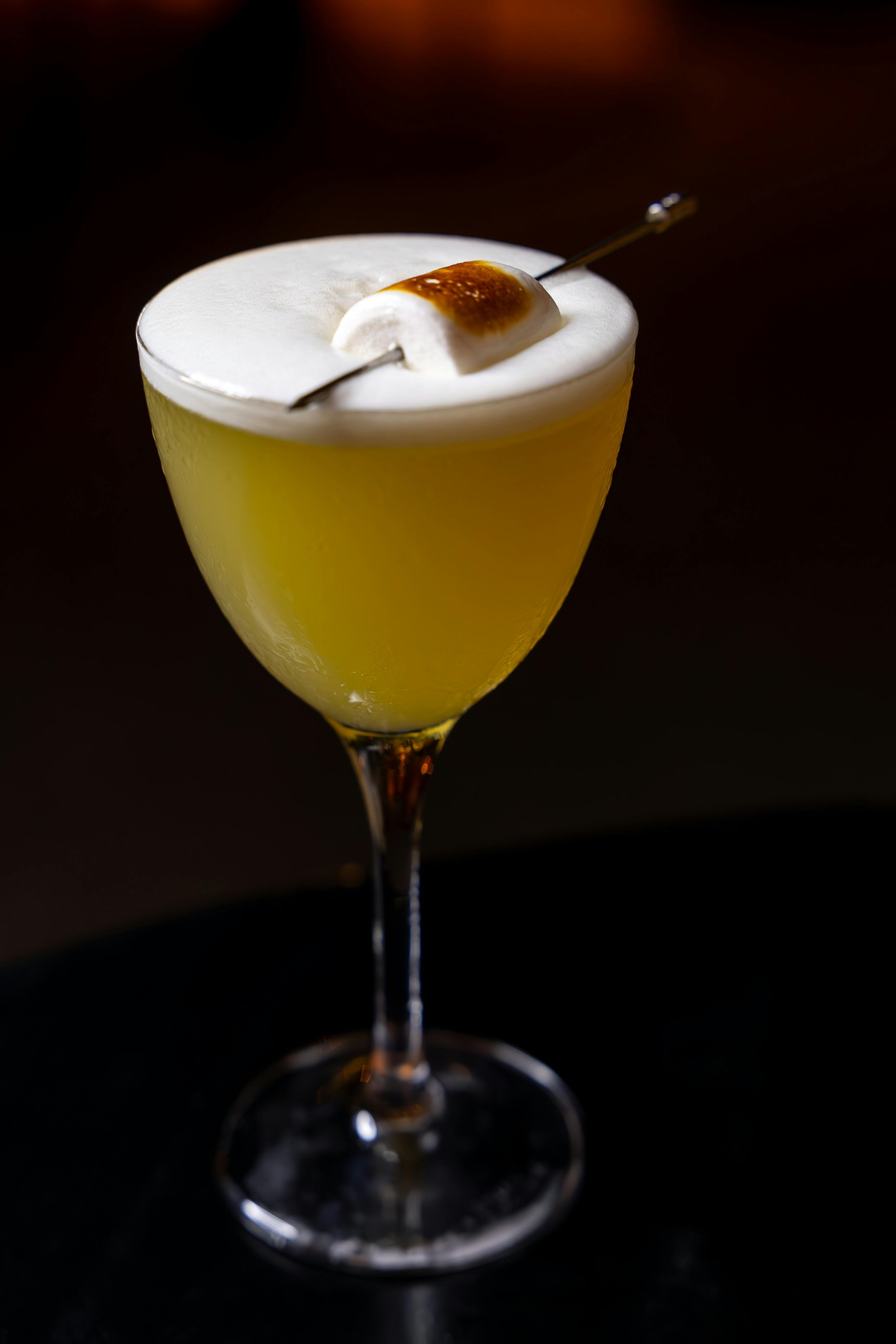 A cocktail in a stemmed glass topped with whipped cream and a toasted marshmallow, set against a black background.
