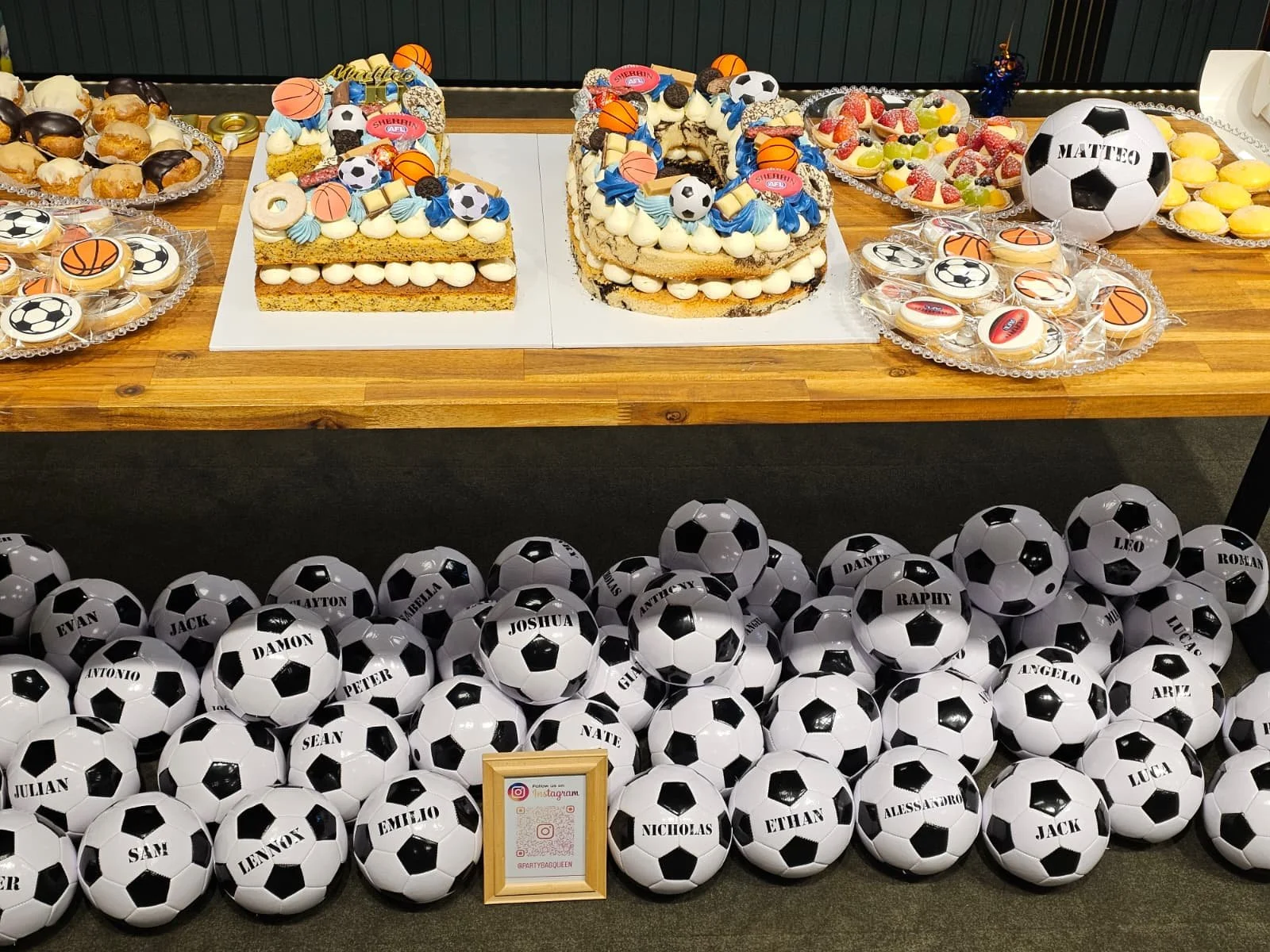 Table decorated with soccer-themed cakes, cupcakes, and candies, surrounded by soccer balls with printed names, likely for a sports-themed celebration.