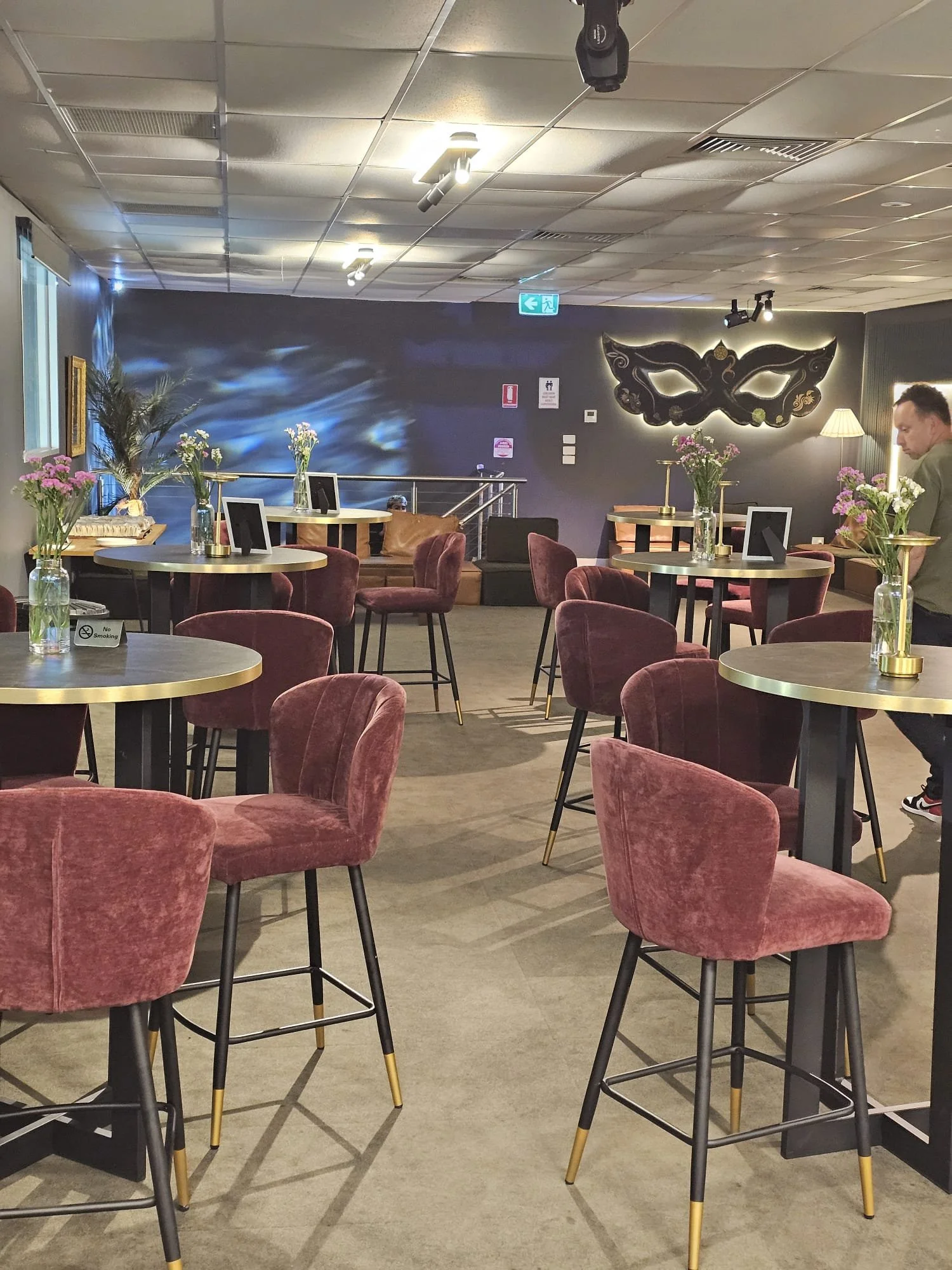 Interior of a modern restaurant or cafe with round tables, pink velvet chairs, and floral centerpieces. There is a wall with a decorative mask, a lamp, and some artwork in the background, along with background seating and emergency exit signs.