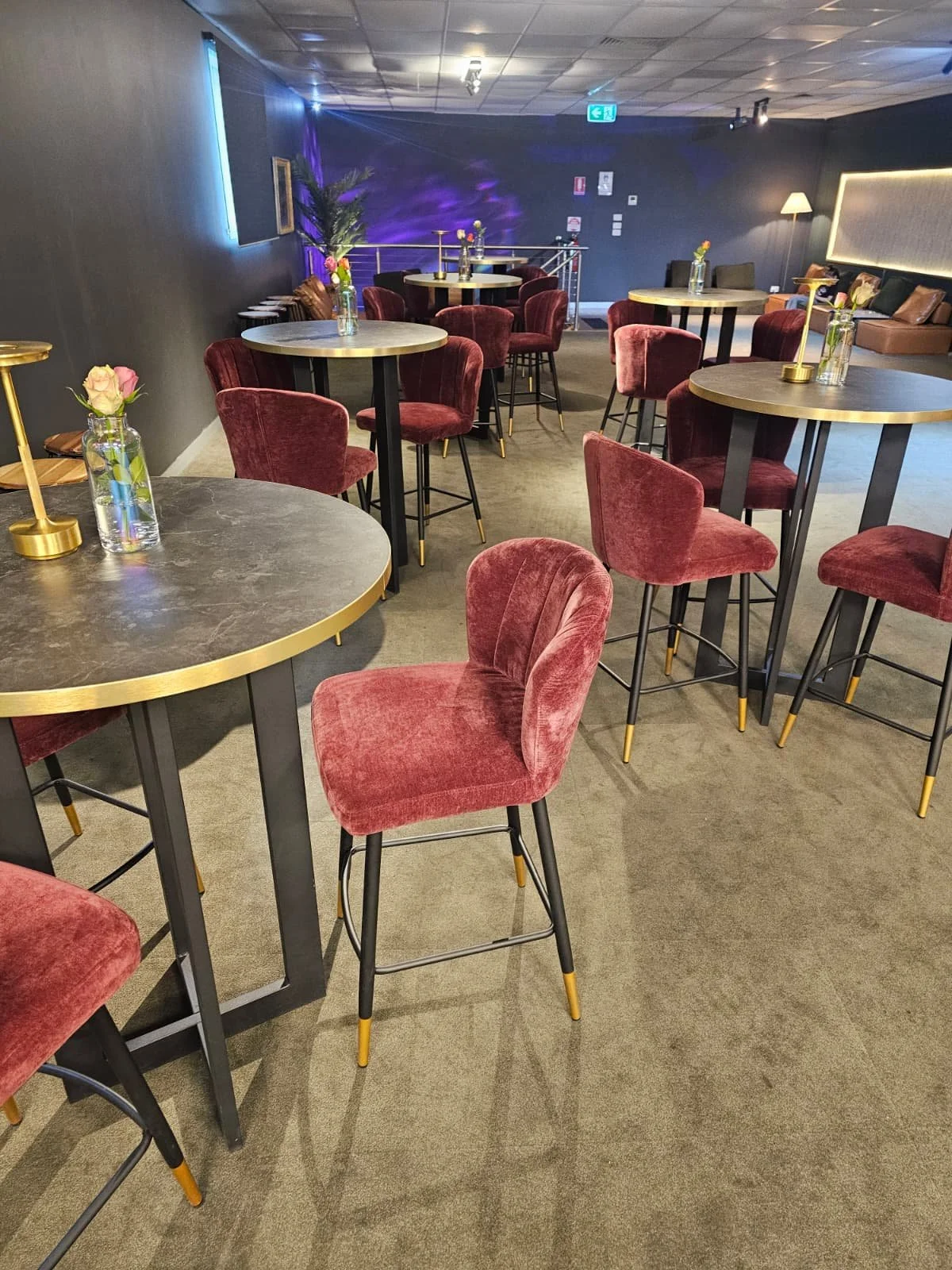 Interior of a modern lounge or cafe with pink velvet chairs, round tables, and small vases of flowers, illuminated by soft lighting.