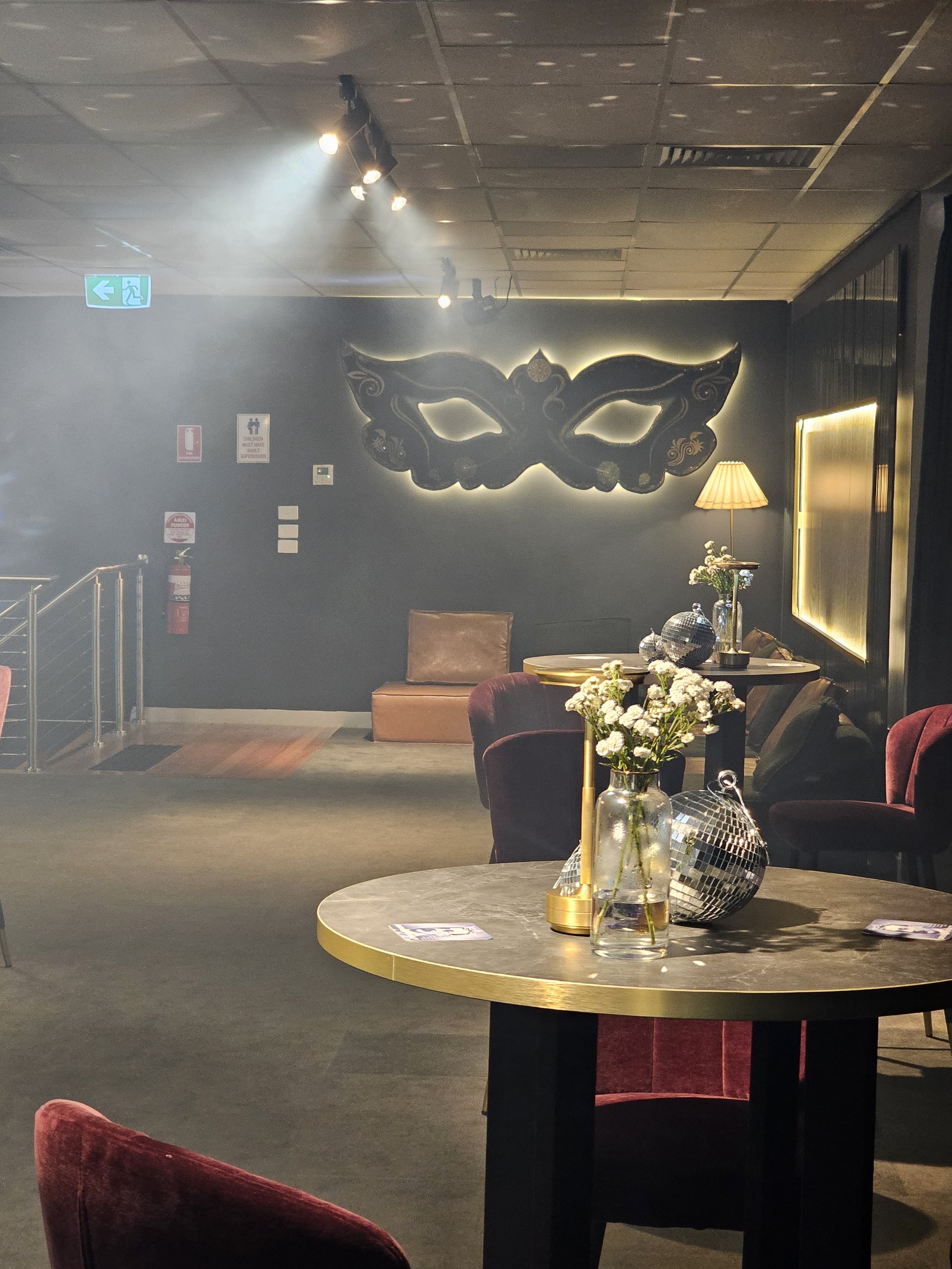 An interior space featuring a black wall with a large decorative butterfly mask, a table with a flower arrangement, a lamp, and a reflective disco ball. There are plush chairs and decorative lighting.